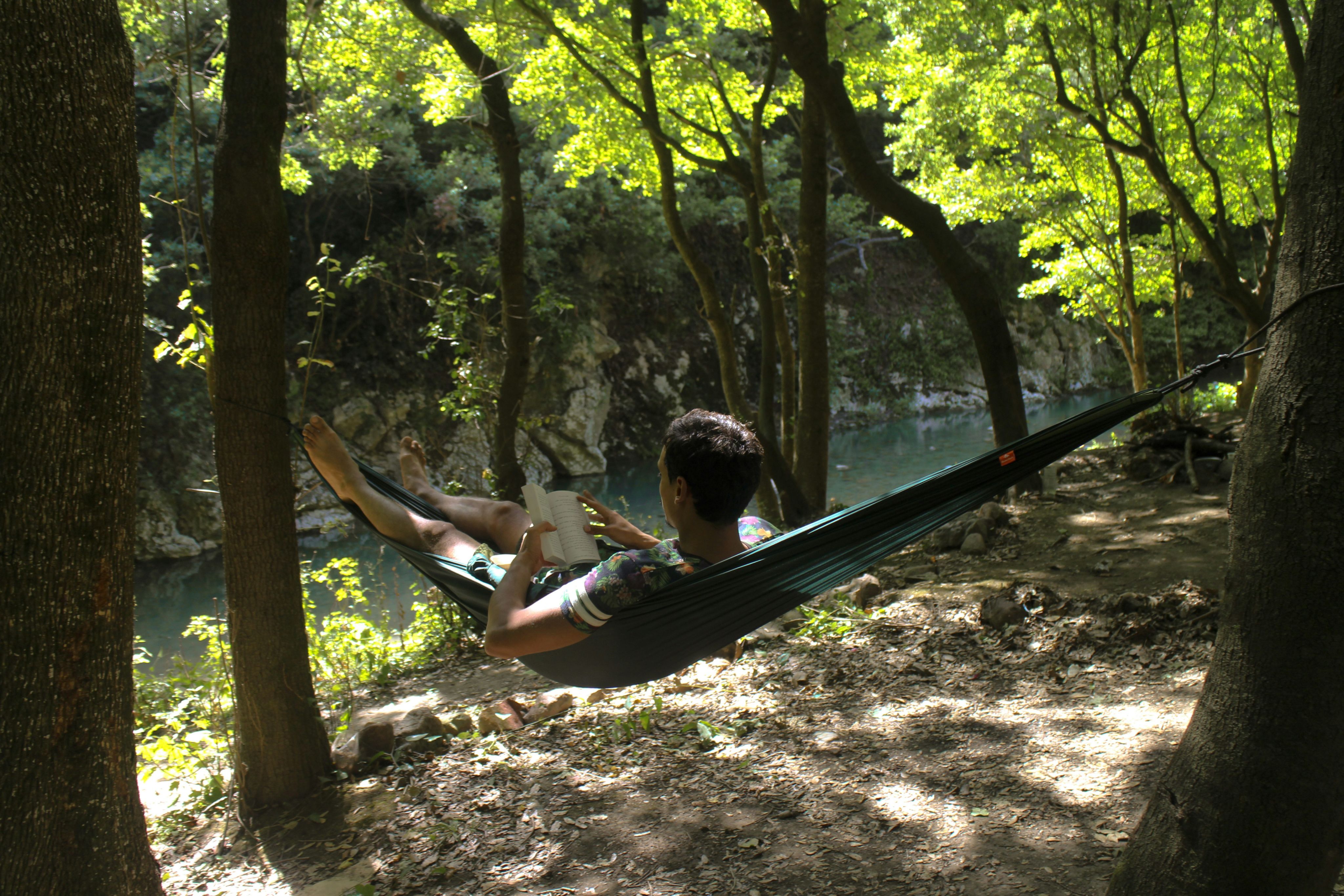man lying down on hammock while reading book near river